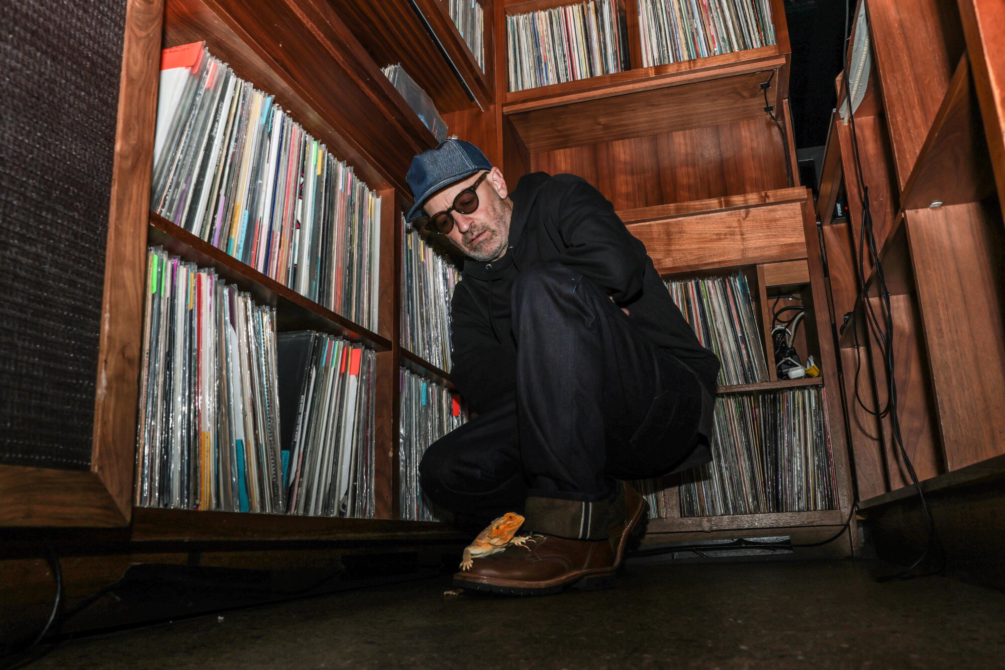 A man in a hat and sunglasses crouches near shelves of vinyl records in a bar.