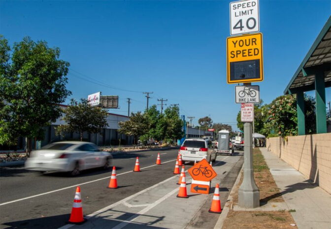 A car passes by orange cones and a yellow speed sign on a street.