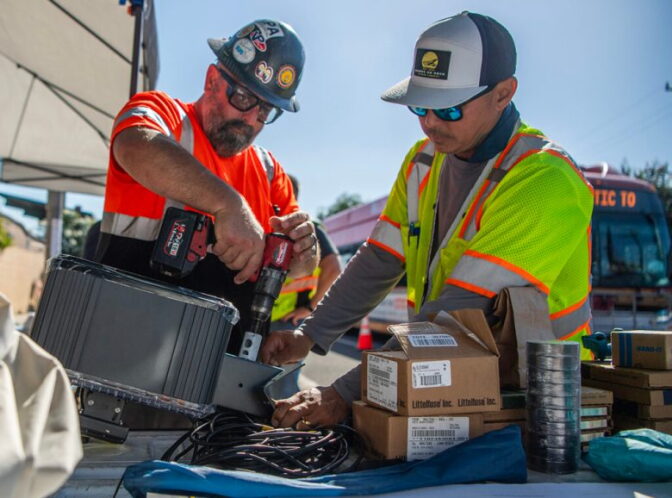 Two men wearing safety vest and shirts use tools on a speed camera with boxes around it on the bed of a pick up truck.