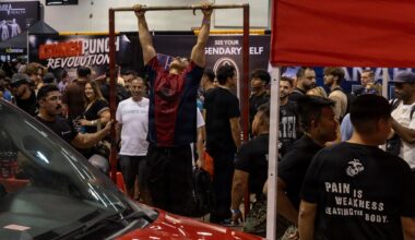 A fitness or health expo event where a person is performing pull-ups on a freestanding pull-up bar surrounded by a large crowd of spectators. Several vendor booths are visible in the background, including