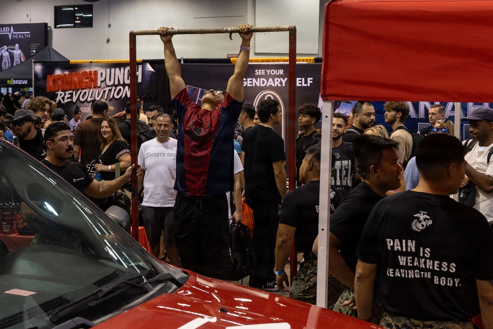 A fitness or health expo event where a person is performing pull-ups on a freestanding pull-up bar surrounded by a large crowd of spectators. Several vendor booths are visible in the background, including