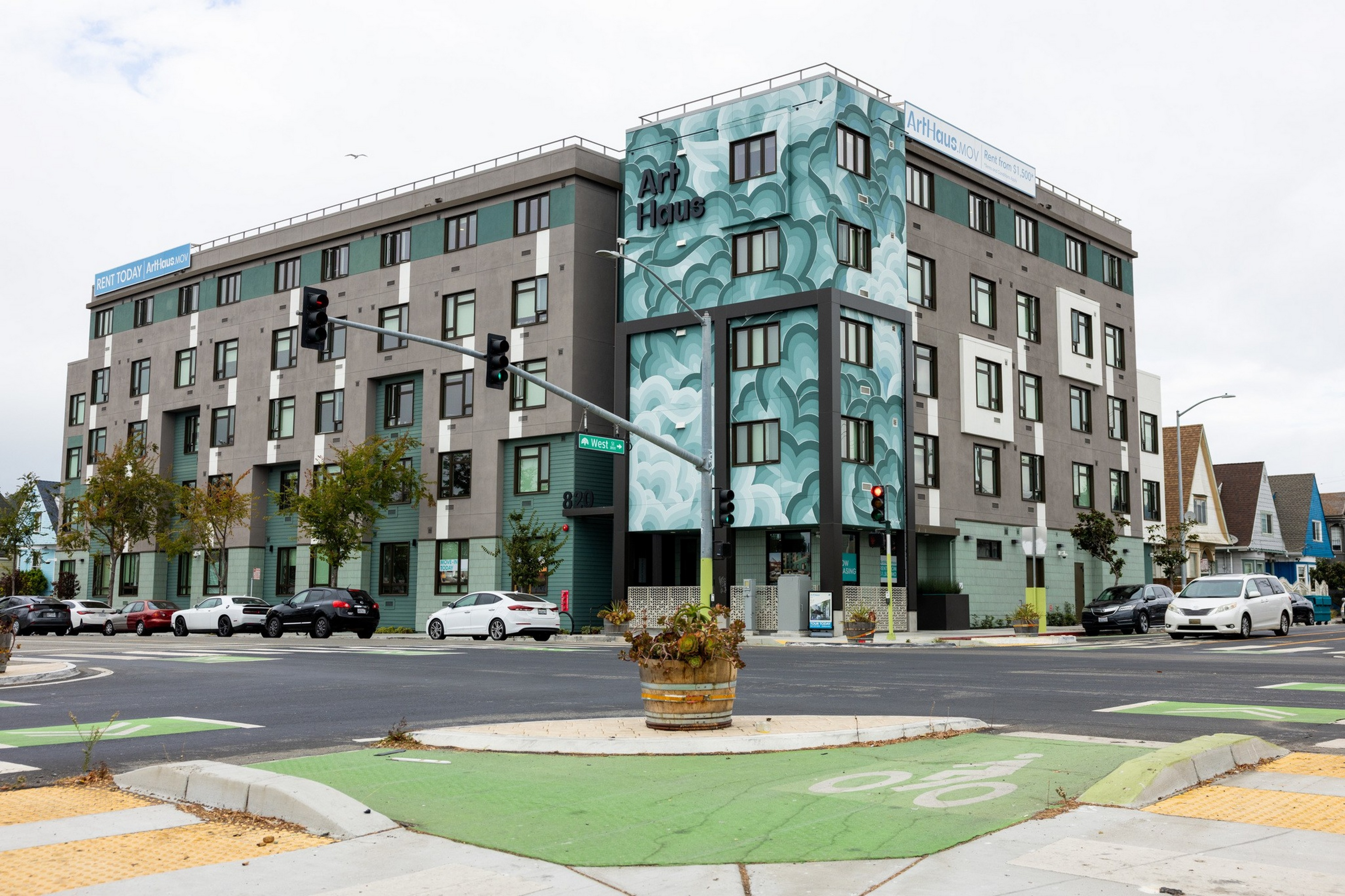 A modern four-story building named “Art Haus” features a teal mural on one corner, with cars parked along the street and traffic lights at the intersection.