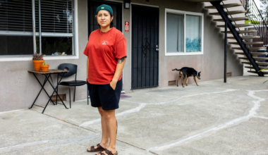 A person in a red shirt, black shorts, sandals, and a green cap stands outside an apartment building, with a dog walking near a window in the background.