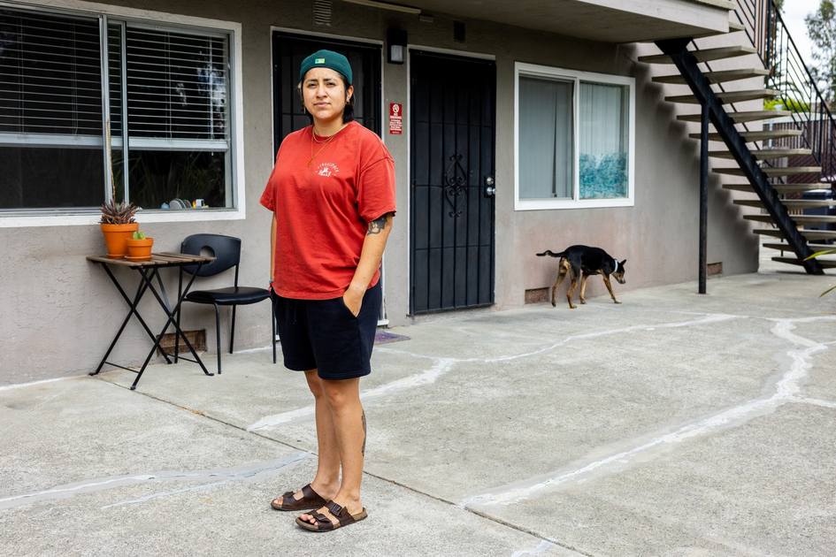 A person in a red shirt, black shorts, sandals, and a green cap stands outside an apartment building, with a dog walking near a window in the background.
