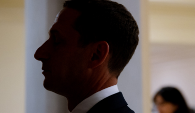 A man in profile with eyes closed wears a dark suit and white shirt, standing against a softly lit background with a blurred figure behind him.