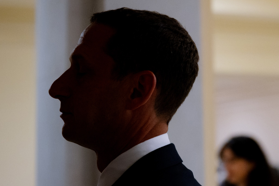 A man in profile with eyes closed wears a dark suit and white shirt, standing against a softly lit background with a blurred figure behind him.