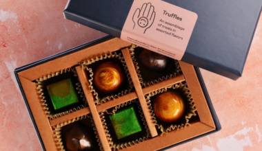 A box contains six assorted chocolates: two green square pieces, two shiny brown spheres, and two dark brown round pieces in paper liners.