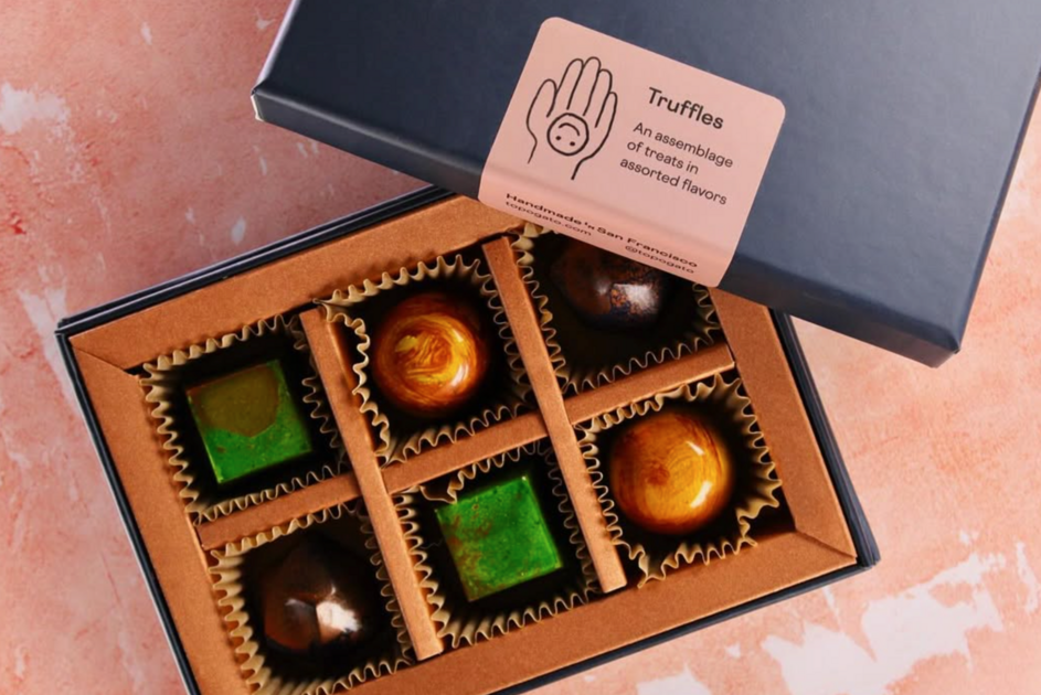 A box contains six assorted chocolates: two green square pieces, two shiny brown spheres, and two dark brown round pieces in paper liners.