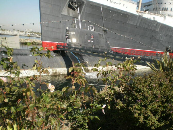 A submarine in shallow water is docked in between a large ship and bushes.