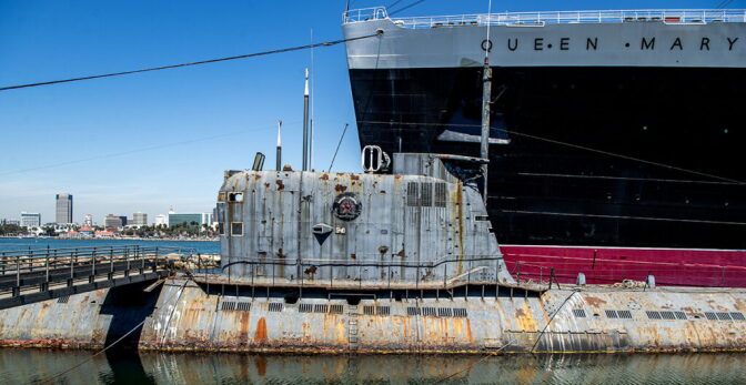 A close up of a rusted submarine docked in shallow water next to a large ship.