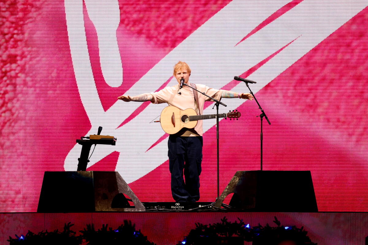 Ed Sheeran holds his arms out while holding a guitar and speaking into a microphone