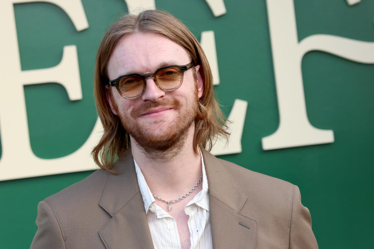  Finneas O'Connell in a beige suit and sunglasses poses agains a green and beige backdrop