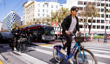 A man in sunglasses rides a blue Lyft bike across a city crosswalk, while two other cyclists and a red bus wait behind him on a sunny day.