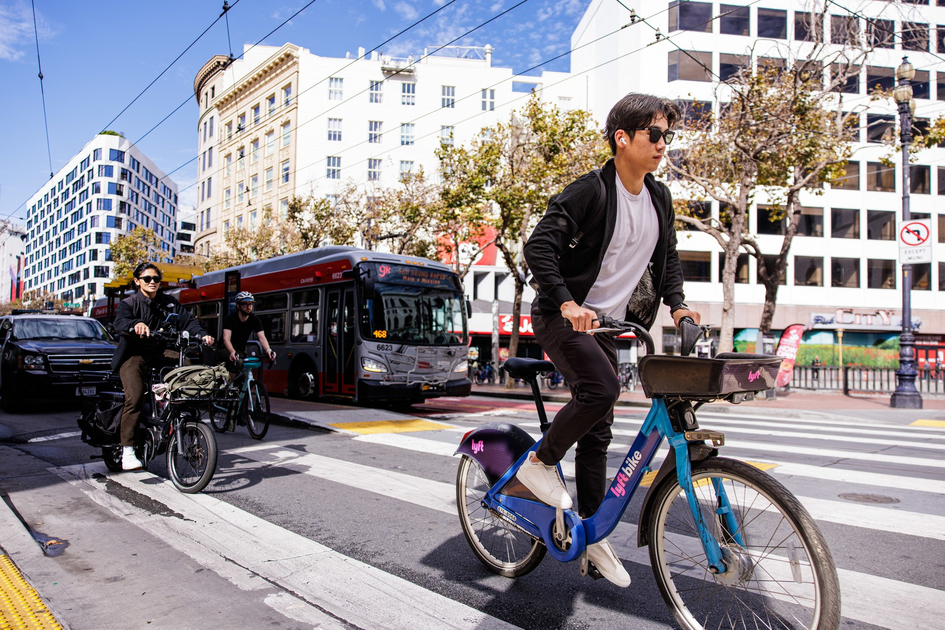 A man in sunglasses rides a blue Lyft bike across a city crosswalk, while two other cyclists and a red bus wait behind him on a sunny day.