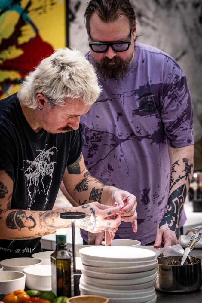 Two chefs with tattooed arms plating food together at a restaurant counter — one light-skinned with short brown hair, a beard, glasses, and a purple graphic shirt; the other light-skinned with short bleached-blond hair and a mustache, plating a dish by hand.