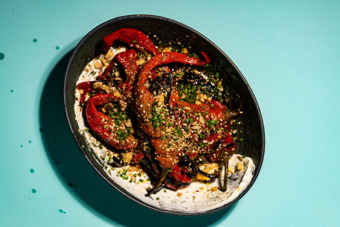 Charred red Jimmy Nardello peppers topped with dukkah and chives, resting on whipped labneh in a dark oval dish against a teal background.