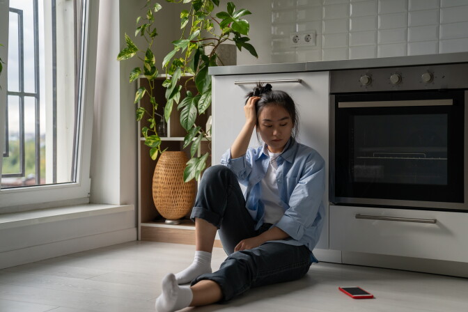 A frustrated female presenting person sits on floor in kitchen looking at smartphone waiting for call or message. She has a medium-light skin tone and wears a white t-shirt with a light blue dress shirt on top, as well as dark jeans and white socks.