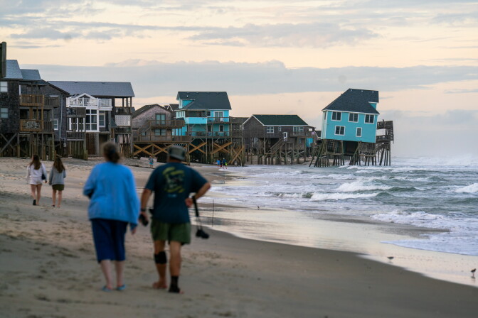 People walking along a beach. Houses on it are collapsing.