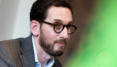 A man with short dark hair, beard, and glasses wears a brown plaid blazer over a light blue shirt, sitting with hands clasped against a dark blue background.