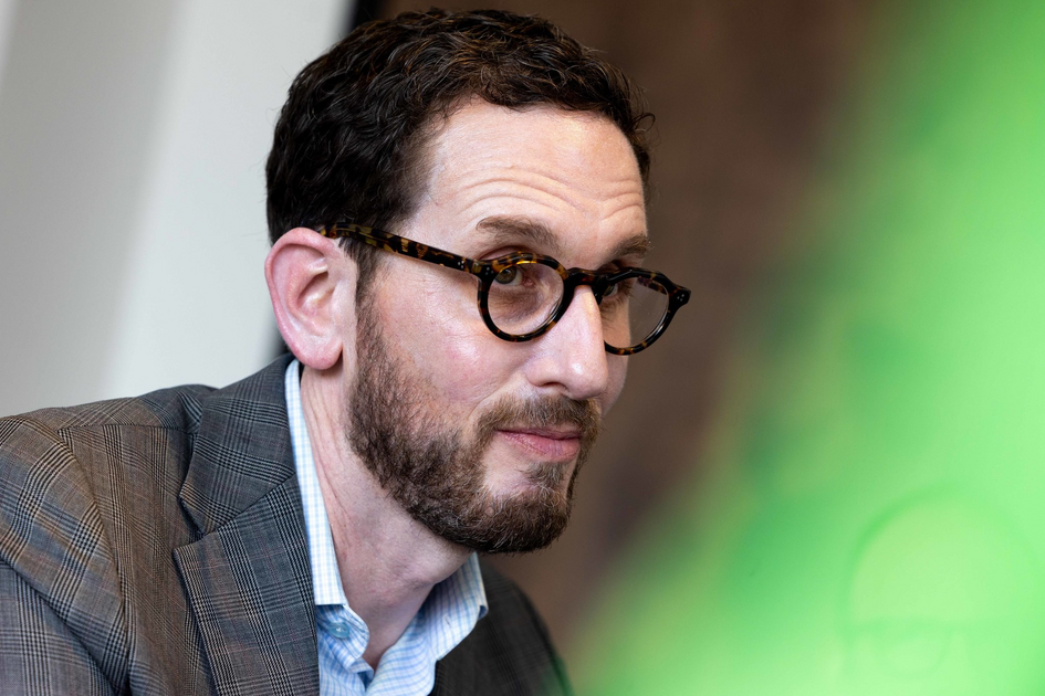 A man with short dark hair, beard, and glasses wears a brown plaid blazer over a light blue shirt, sitting with hands clasped against a dark blue background.
