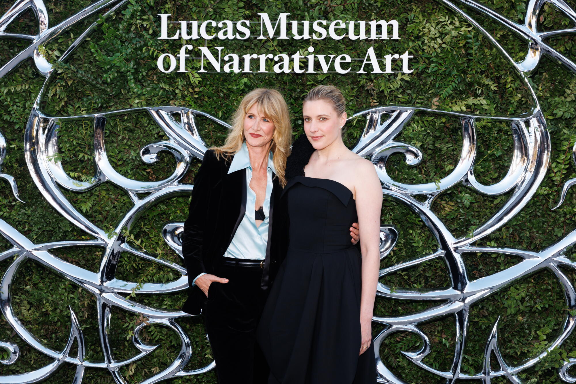 Laura Dern and Greta Gerwig stand in front of a grass wall.