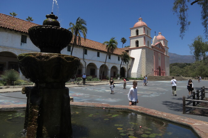 A water fountain can be seen in the foreground, with the Santa Barbara mission in the background. Numerous people can be seen walking about.
