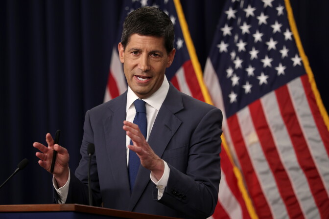 Federal Reserve Chair Kevin Warsh speaks at a podium during a news conference with a dark grey suit and navy tie. Two American flags can be seen behind him.
