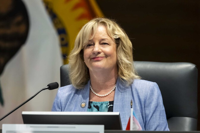 A middle-aged woman with short blond hair and light skin tone wearing a light blue blazer and pearls sits a dais with a microphone pointed towards her. She has a slight smile on her face.