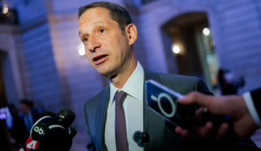 A man in a suit and tie is speaking to reporters holding microphones and a smartphone near a building lit with blue light.