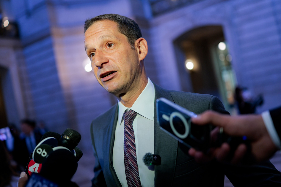 A man in a suit and tie is speaking to reporters holding microphones and a smartphone near a building lit with blue light.