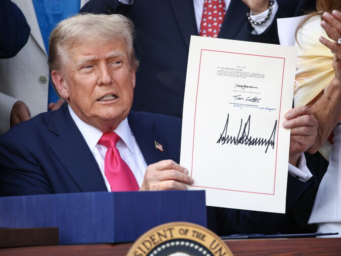 A light-skinned man with blond hair holds up a signed document with a large signature at a podium bearing a presidential seal, surrounded by other people.