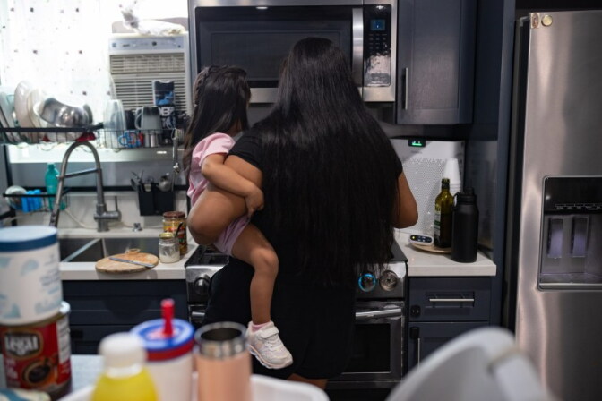 A mother holds up her daughter in a kitchen facing away towards a stove.