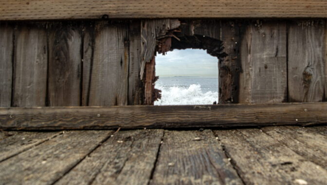 A low angle view looking through a hole of a seawall on a boardwalk seeing waves crashing from the ocean.