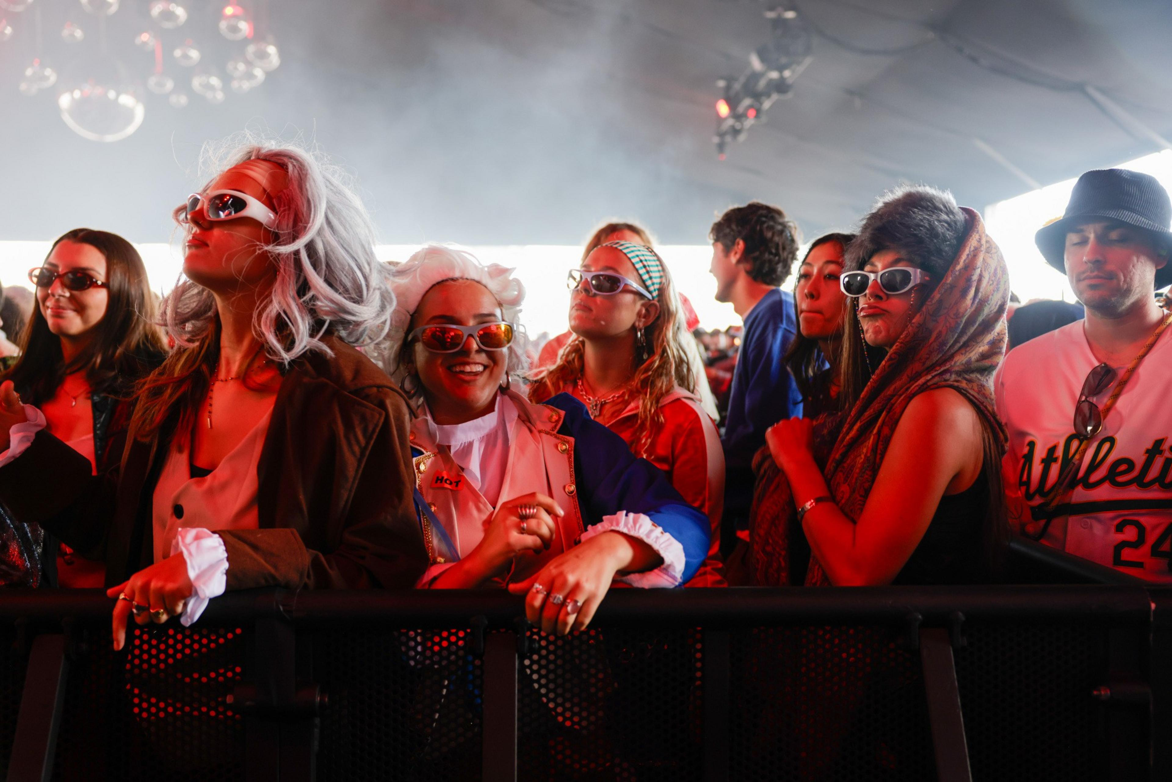 A group of young people wearing sunglasses and colorful outfits stand closely behind a metal barrier, enjoying an indoor event with red lighting.