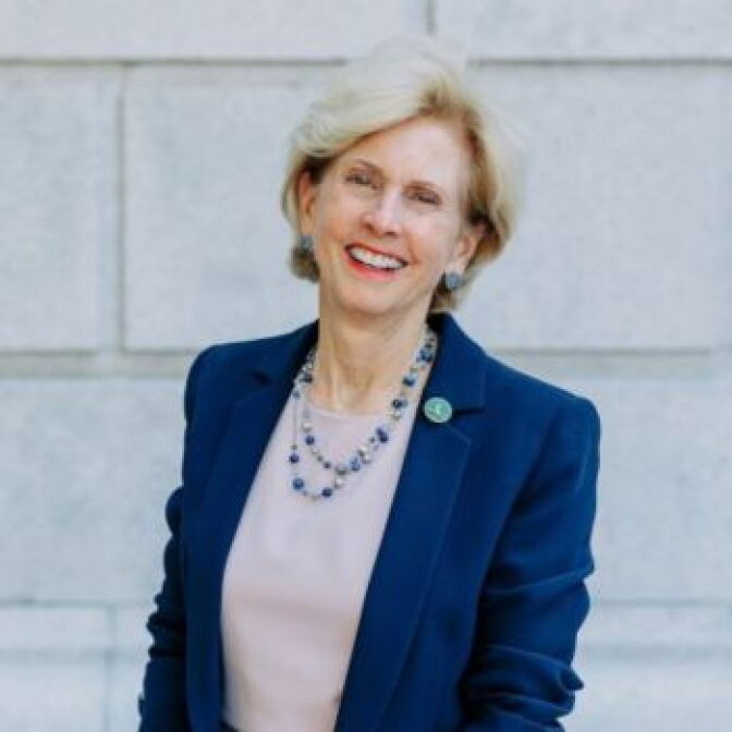 A woman with a short, blonde bob is laughing for the camera. She is wearing a blue jacket over a pale-colored dress.