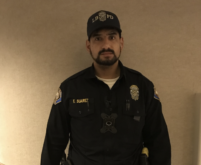 A uniformed man with medium skin tone has a ballcap with LBPD, a badge and E Suarez on his shirt.