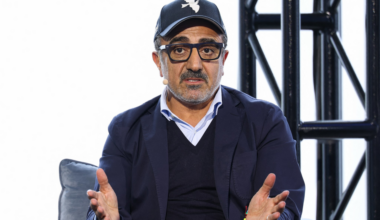 A man wearing glasses and a black cap with a white dove emblem gestures with both hands while speaking, seated on a gray chair.