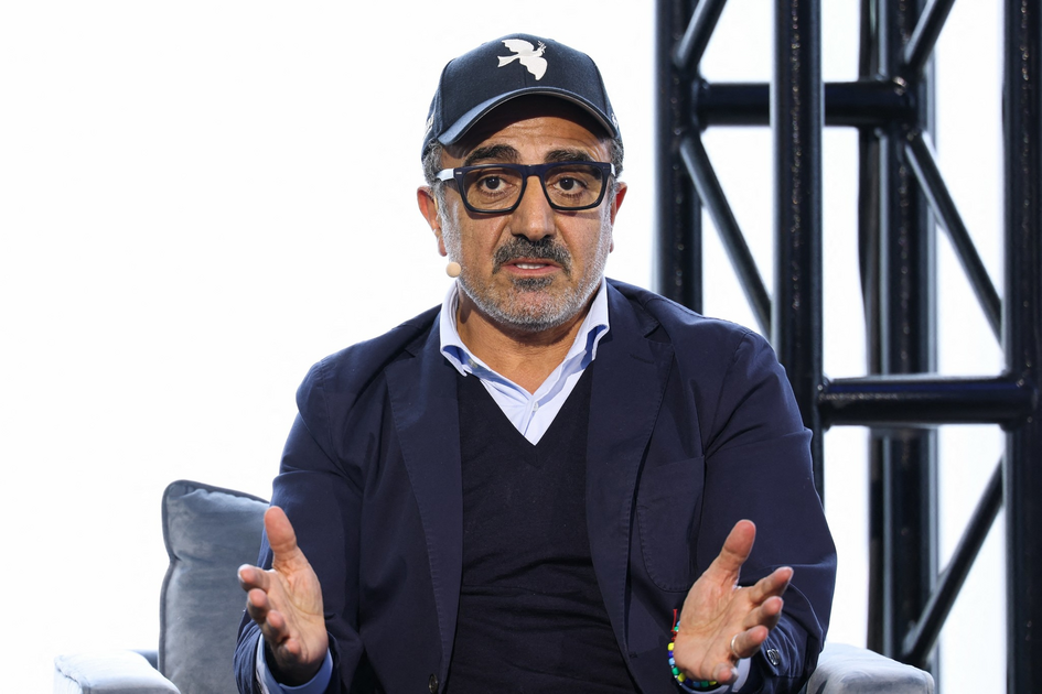 A man wearing glasses and a black cap with a white dove emblem gestures with both hands while speaking, seated on a gray chair.