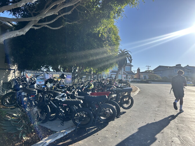 Rows of electric bikes with fat tires line a fence under a tree as a teenager walks down the street in sunlight.