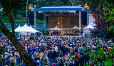 A band performs on a large outdoor stage with a crowd gathered on a grassy area. The backdrop features a bridge design, and trees surround the venue.