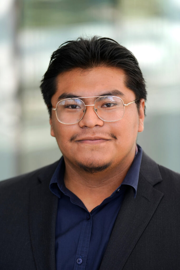 Headshot of Israel Sanchez in a dark suit jacket and blue button-down shirt wearing wire-rimmed glasses in front of a fuzzy green background