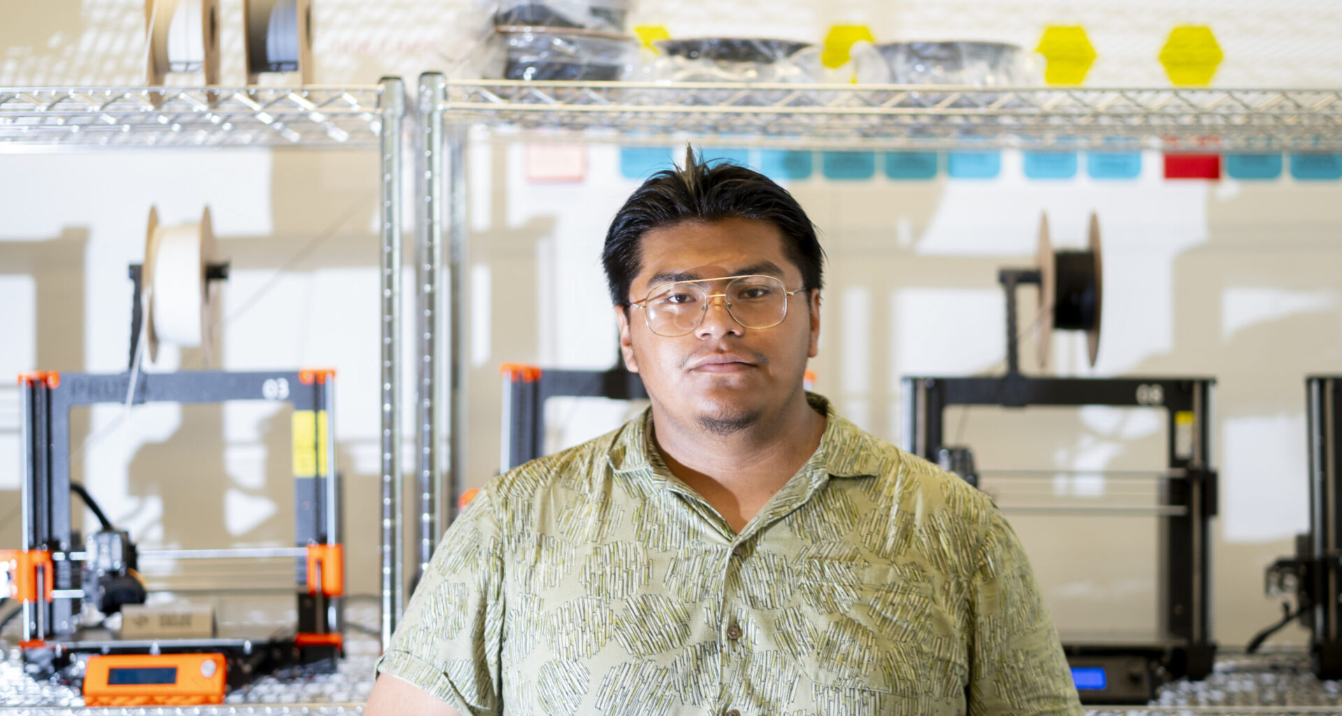 Portrait of Israel Sanchz standing in front of wire shelves of engineering equipment wearing a light green short-sleeve button-down shirt and wire-rimmed glasses