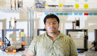 Portrait of Israel Sanchz standing in front of wire shelves of engineering equipment wearing a light green short-sleeve button-down shirt and wire-rimmed glasses