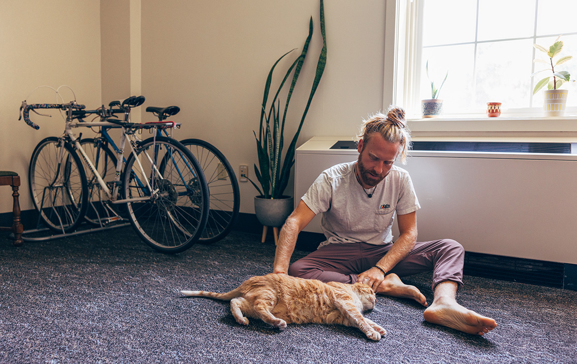 A man pets his cat in a carpeted dorm room, surrounded by bikes and plants.