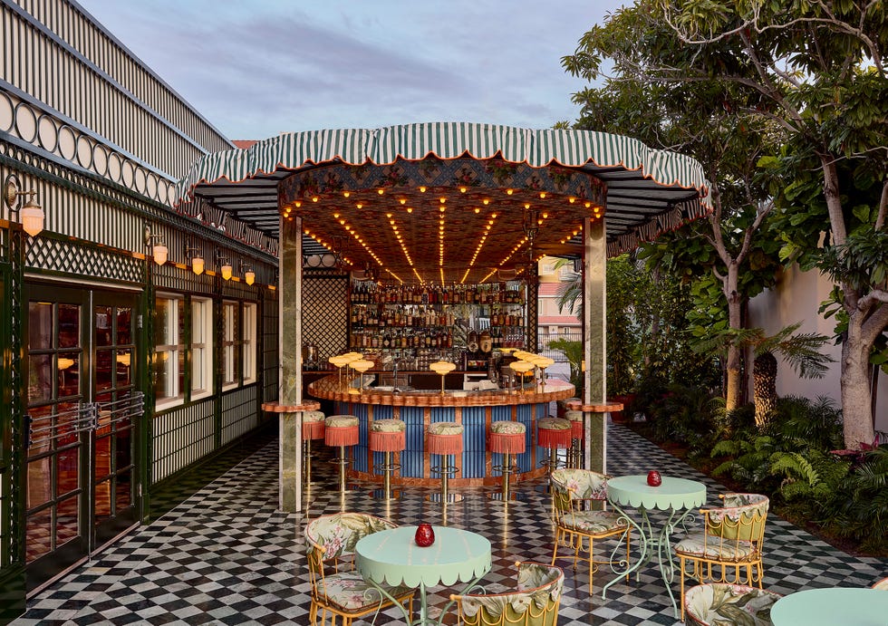 Colorful outdoor bar area with patterned seating and lush greenery, illuminated by warm lights against a twilight sky.