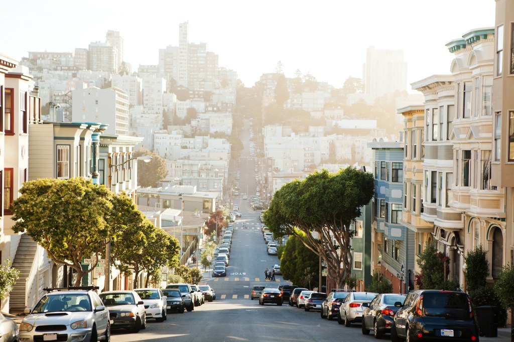 The view from the top of a San Francisco hill looking down a residential street lined with parked cars and multi-colored houses.