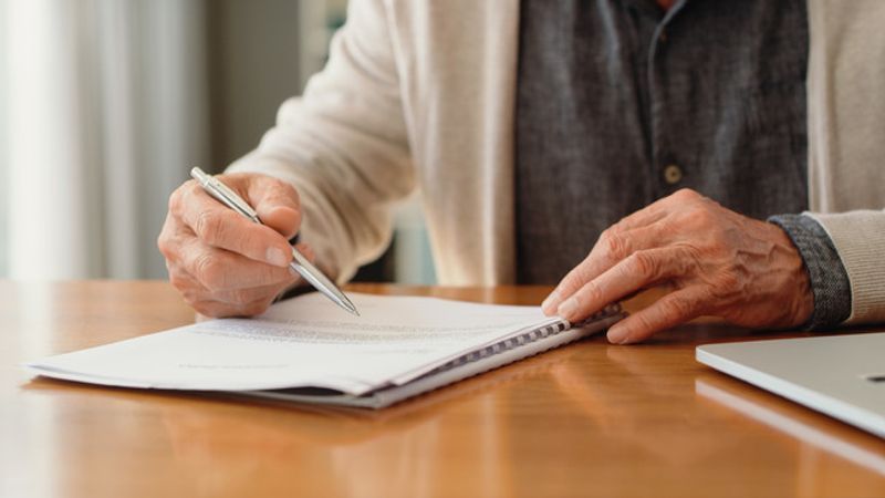 Hands of pension man reading retirement paperwork about policy, investment options and funeral cover while planning finance, insurance and budget at home. Signing will, legal document and contract