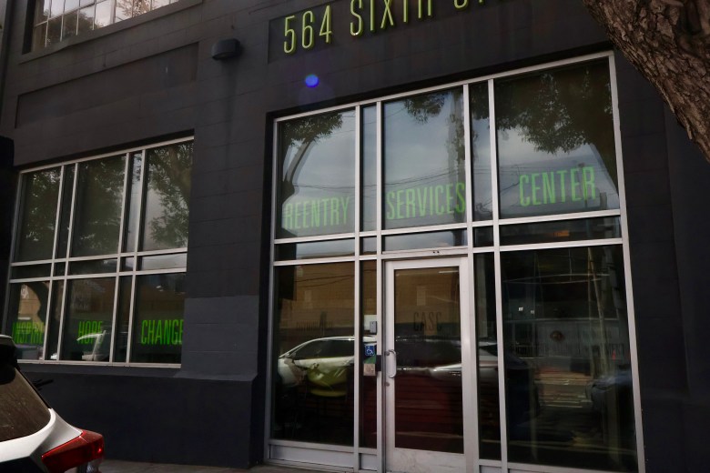Street view of a dark building with large windows displaying “Reentry Services Center” in green letters, located at 564 Sixth St. A car is parked in front, and trees are reflected in the glass. This SF Pretrial facility offers critical support for individuals reentering the community.