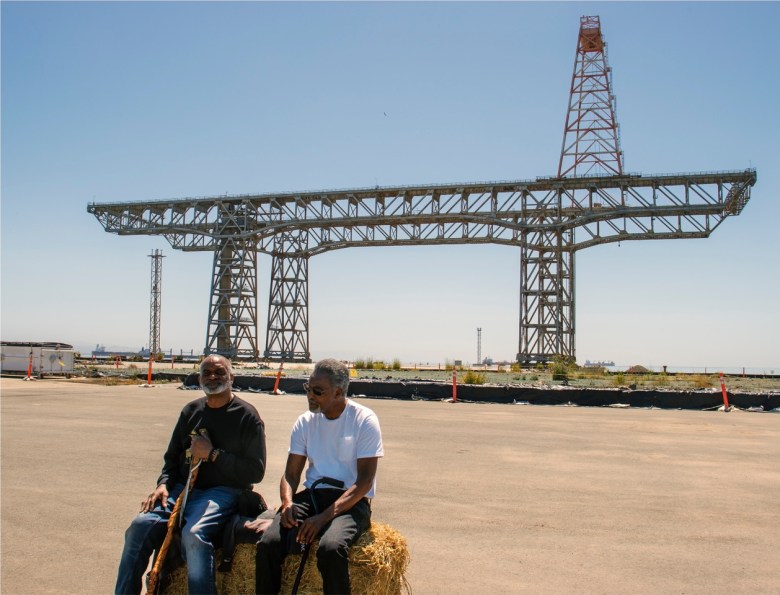 Two men sit on hay bales in front of a large metal industrial structure outdoors on a clear day.