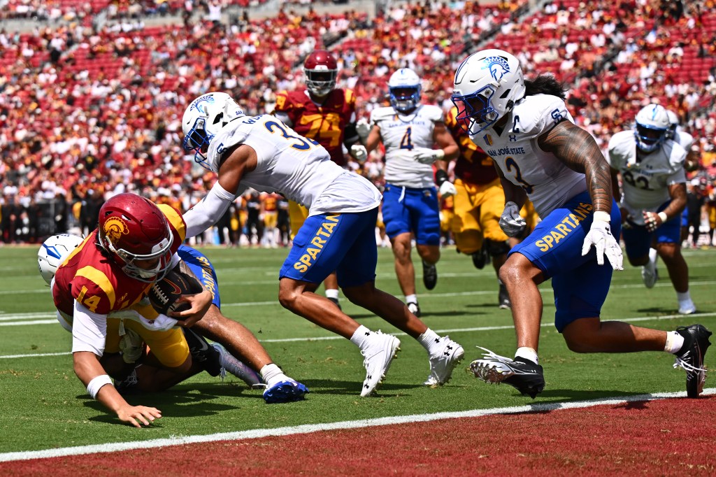 Football player Jayden Maiava, number 14, in a red and yellow uniform, is tackled by an opposing player in a white and blue uniform.